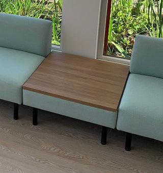 Light blue sofa with a wooden table extension in a room with windows showing greenery outside.