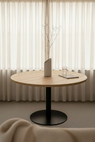 Round wooden table with a black base in front of a sheer curtain
