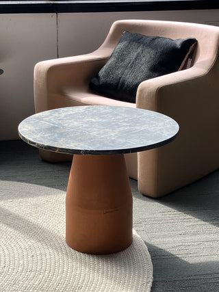 Round marble table with a terracotta cylindrical base in front of a beige armchair with a dark cushion.